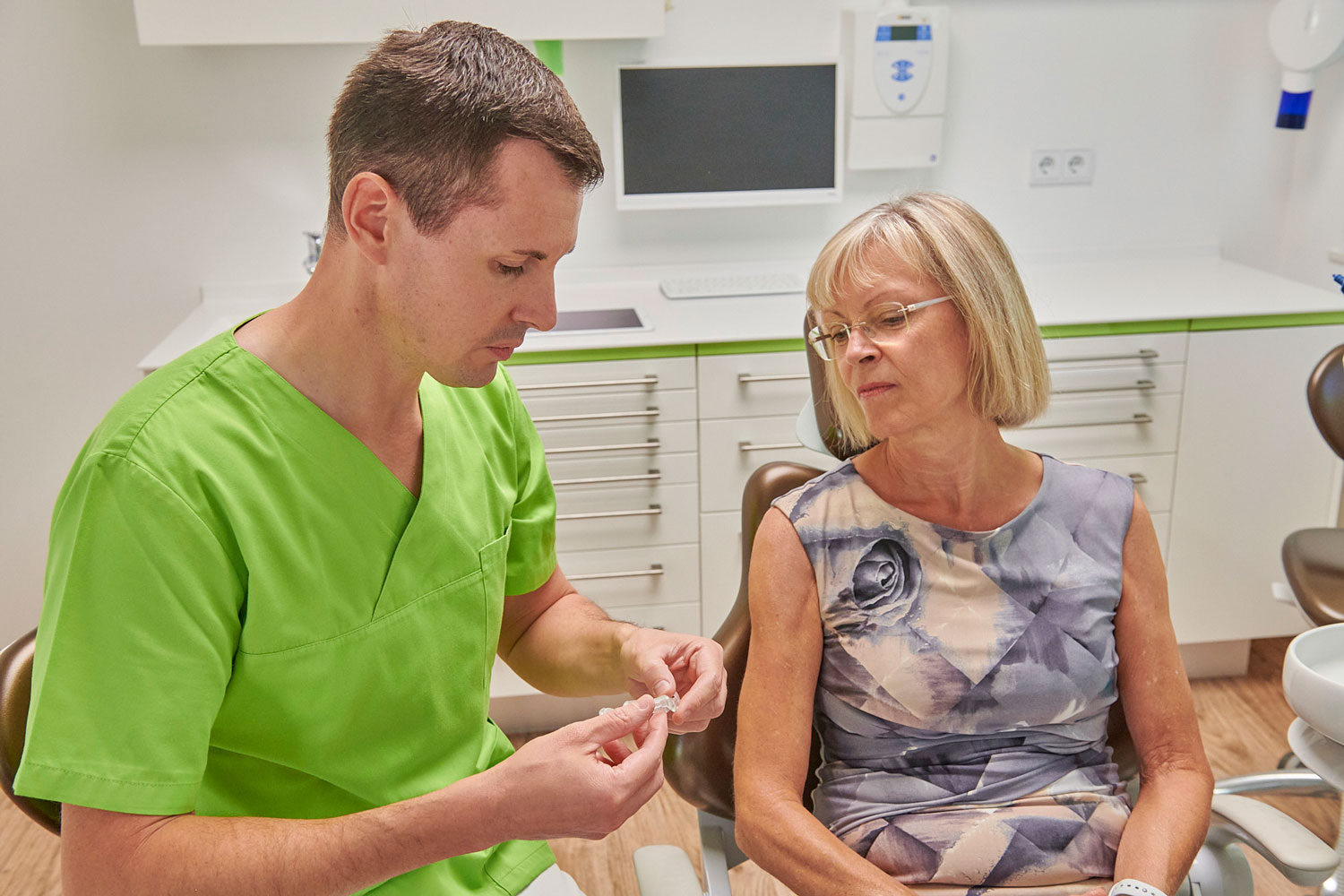 Eine zahnmedizinische Fachkraft in grüner Kleidung erklärt einer älteren Patientin ein kleines Implantat-Modell. Beide sitzen in einem modernen Behandlungsraum.